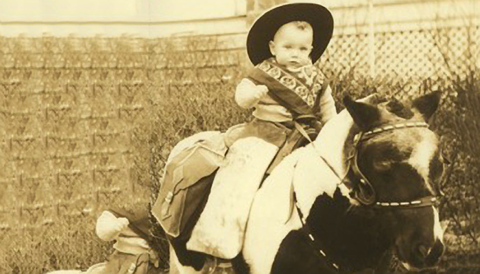 A baby sits on a horse, dressed as a cowboy