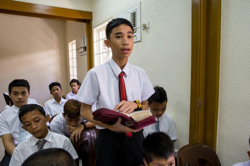 A young man in a white shirt and tie stands up and reads to the class from his scriptures.