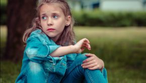 A young girl sits beneath a tree on the grass wearing jeans and a jean jacket with a sad/forlorn look on her face