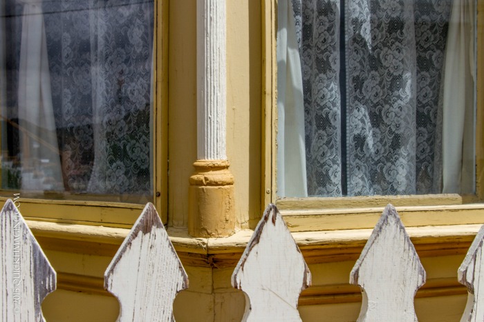 Partial view of picket fence and corner windows covered in lace curtains