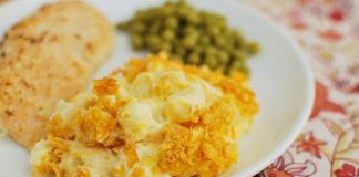 A plate with funeral potatoes, peas and grilled chicken is shown with the potatoes in focus and the rest blurred in the background.