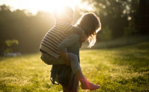 A young girl is giving a little boy a piggyback ride in a grassy area with the sun's glare peaking above their silhouetted heads.