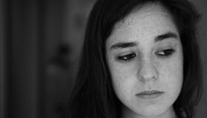 A close up picture of a teenage girl with freckles and long hair. The image is in gray scale and she appears sad