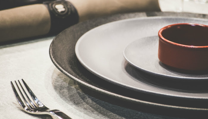 A zoomed in picture of a silver fork sitting beside a fancy place setting of plates and a napkin sits in the background with a fancy napkin ring around it