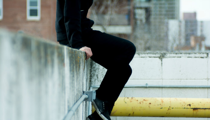 A teenager (gender unclear) dressed all in black is sitting atop of a wall, presumably on a roof top. Their head is cut off and we only see their legs and pipes and outlets on the wall