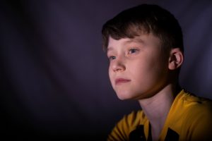 A young Caucasian boy in a yellow shirt is shown from the shoulders up staring off with a calm/almost sad look on his face.