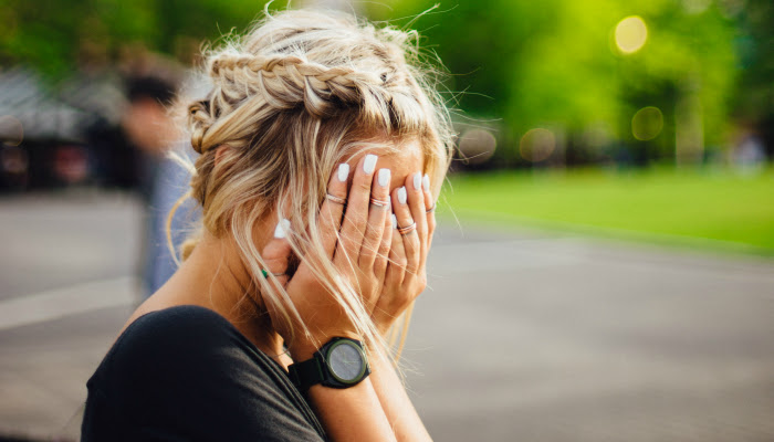 A girl is seen from the shoulders up standing profile with her hands covering her face (we see that there are various rings on her fingers).