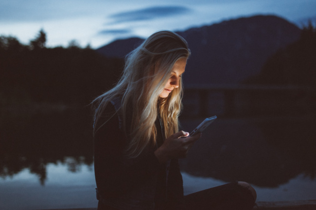 young woman holding phone in the darkness light on her face by a lake